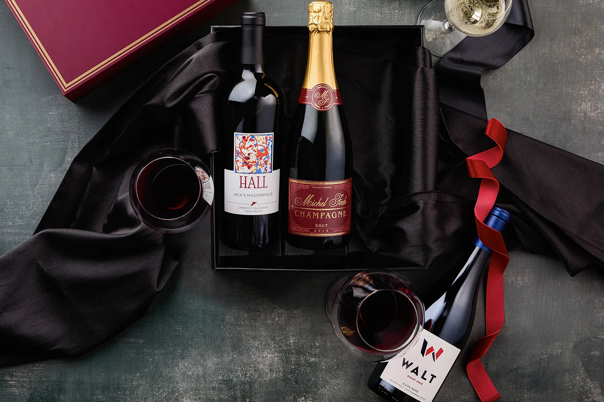 A luxury wine gift set arranged in a black presentation box on a dark textured surface. The set includes a bottle of Hall wine, a bottle of Marie & Louis champagne, a bottle of Walt wine, two filled wine glasses, a red ribbon, and a dark red gift box.