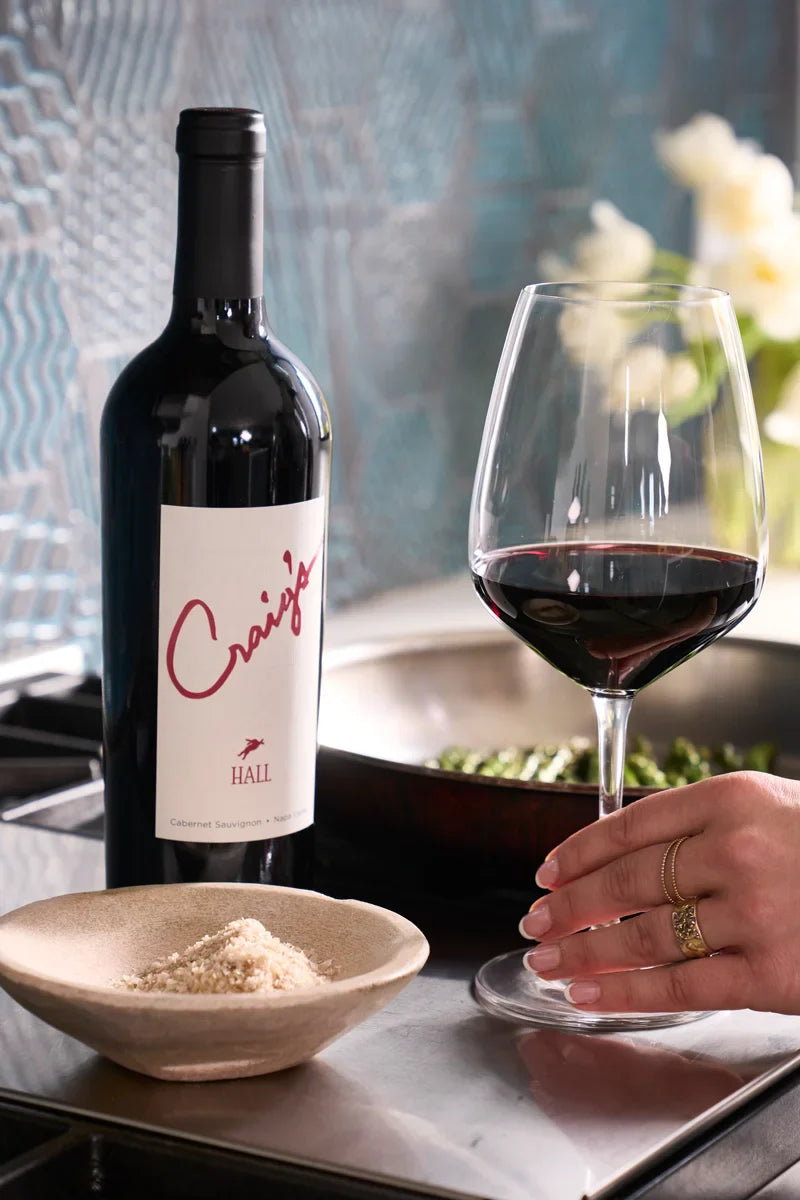 Woman reaching for a glass of red wine next to a bottle of 2023 HALL Craig's Cabernet Sauvignon, with a dish of shredded Parmesan and sautéing vegetables on the stove nearby