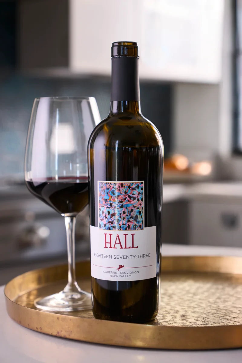 Bottle of 2020 HALL Eighteen Seventy-Three Cabernet Sauvignon on a tray next to a glass of red wine